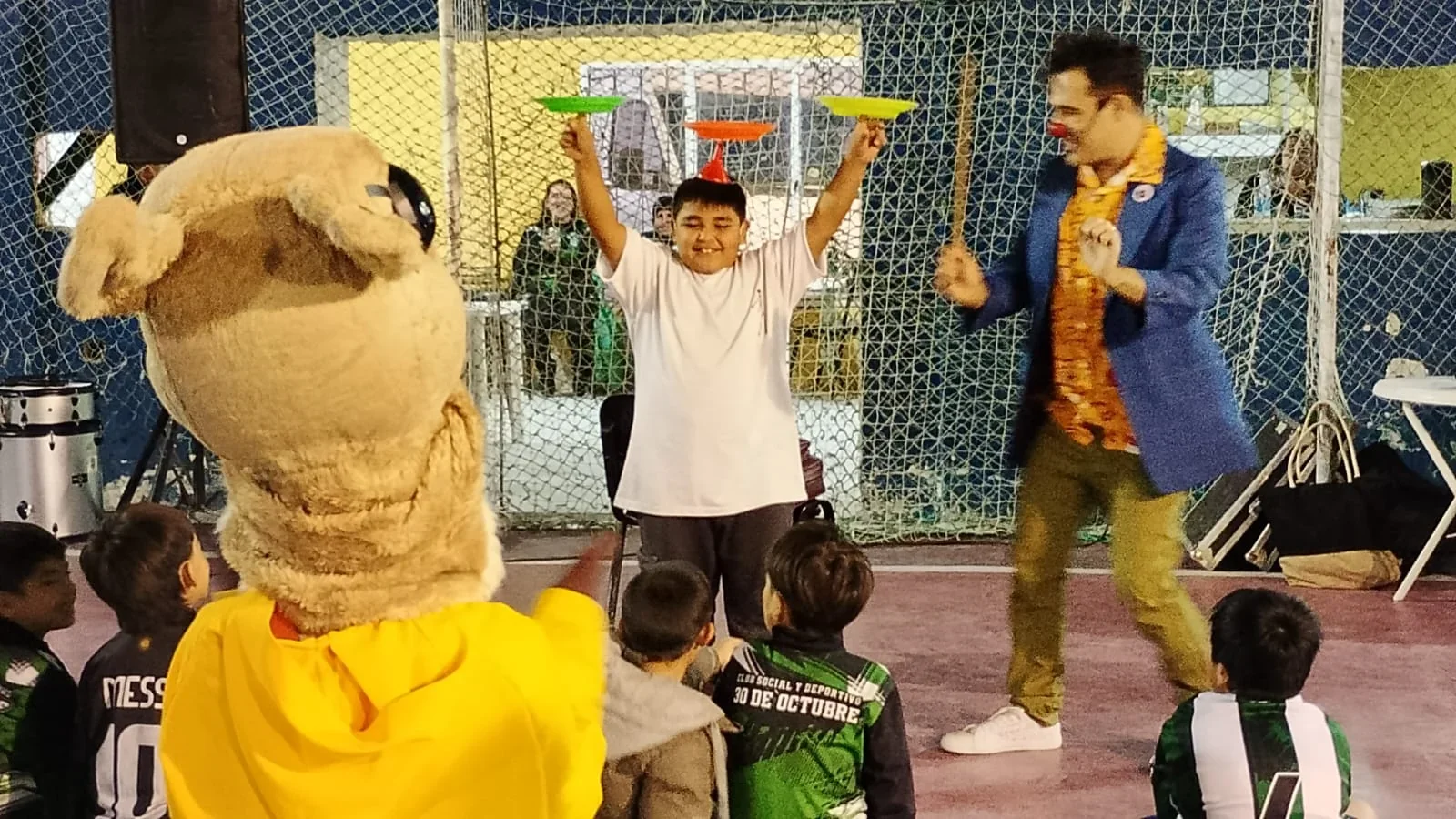 Show de malabares y payaso frente a niños y una mascota en un evento de cultura comunitaria en Comodoro Rivadavia.