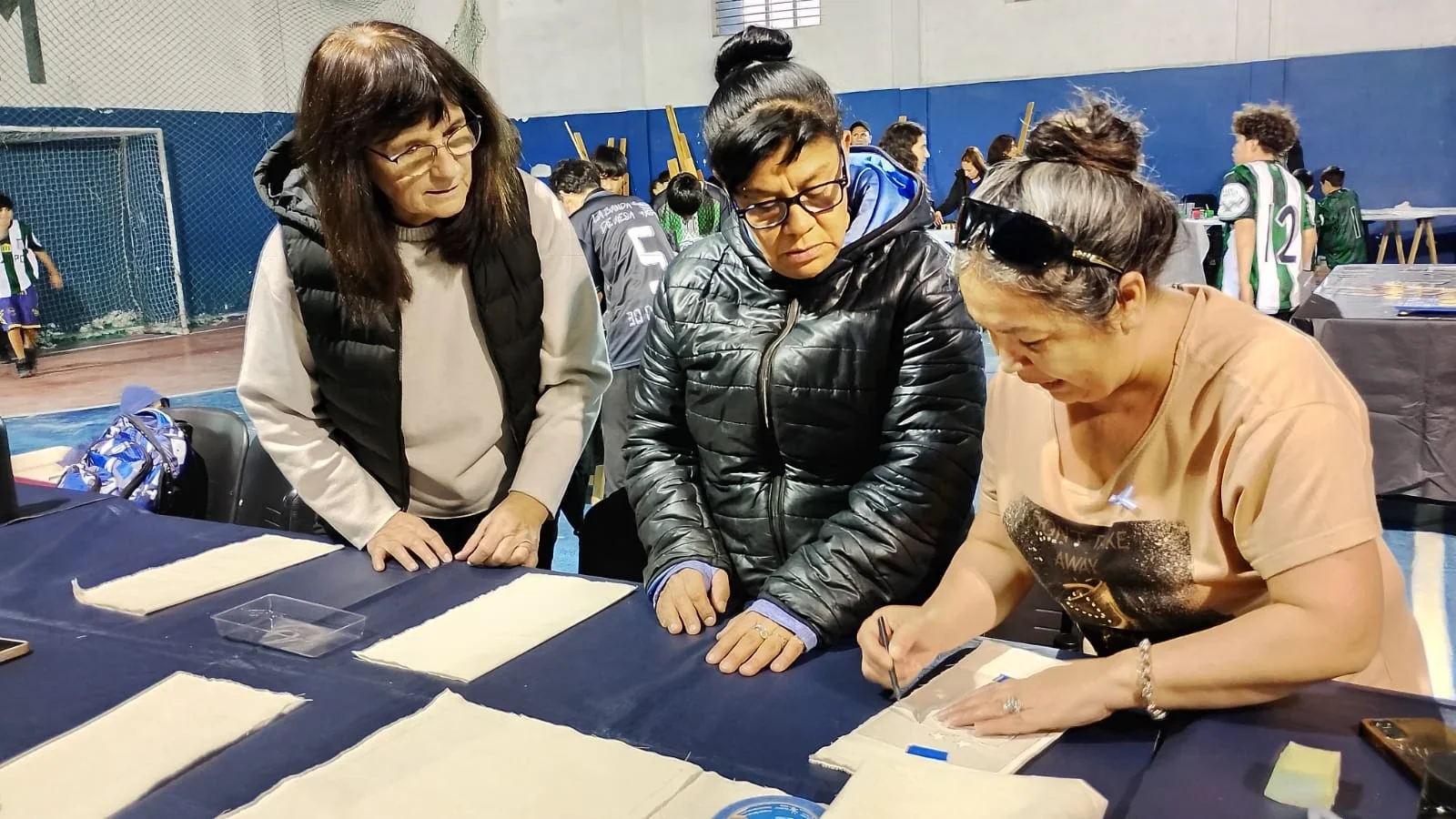 Tres mujeres observan y trabajan sobre papeles en una mesa durante una actividad cultural en Comodoro Rivadavia.