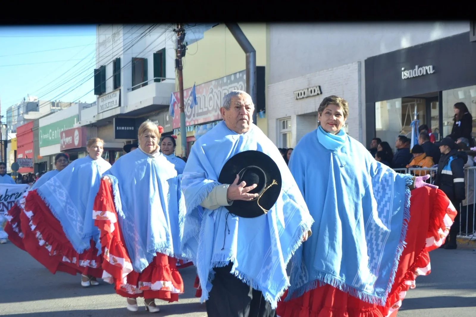 Personas con trajes típicos coloniales, ponchos celestes y faldas rojas, en un desfile cultural en Comodoro Rivadavia.