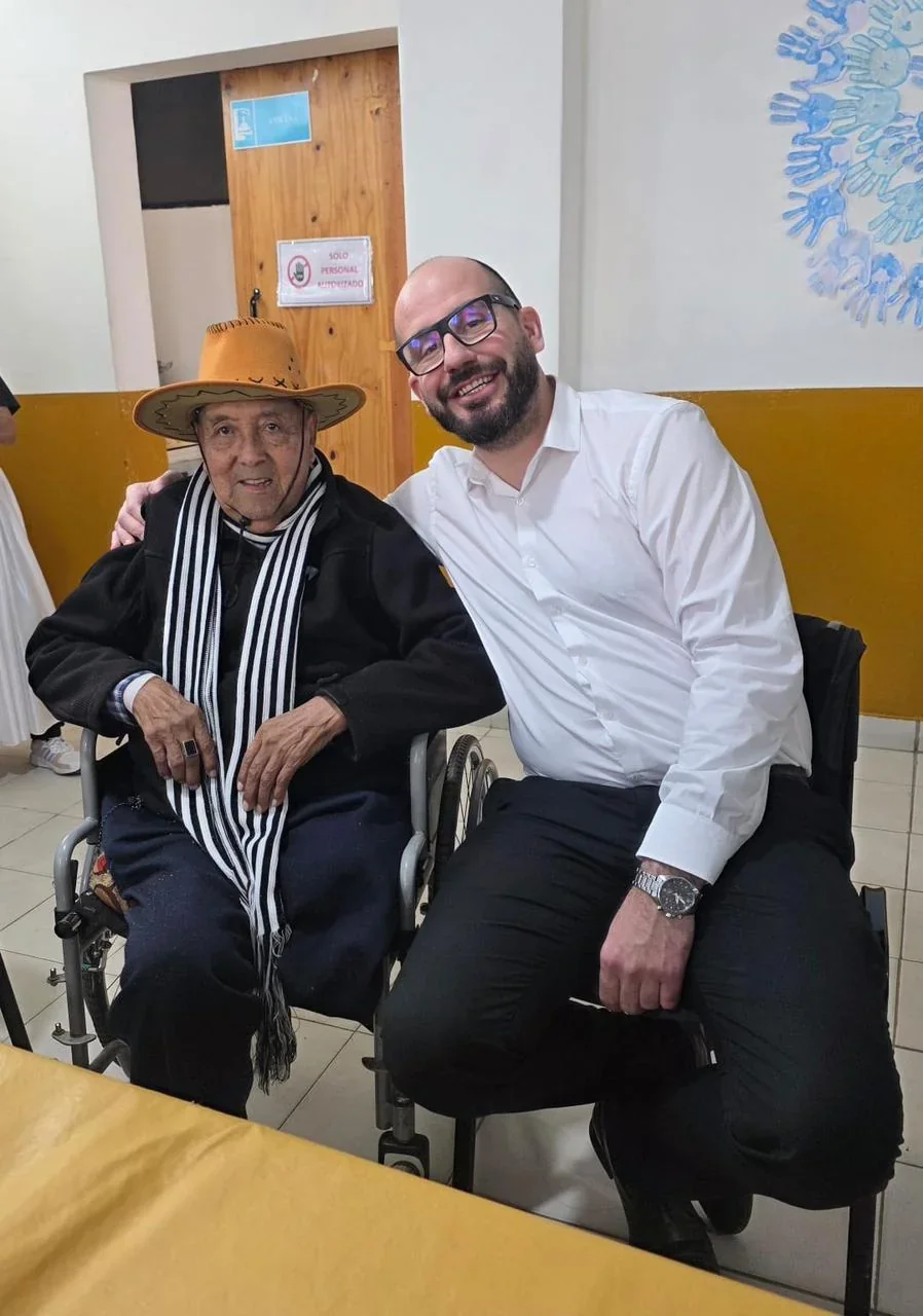 Hombre mayor en silla de ruedas con sombrero y poncho junto a un hombre joven en el Centro de Día Papa Francisco.