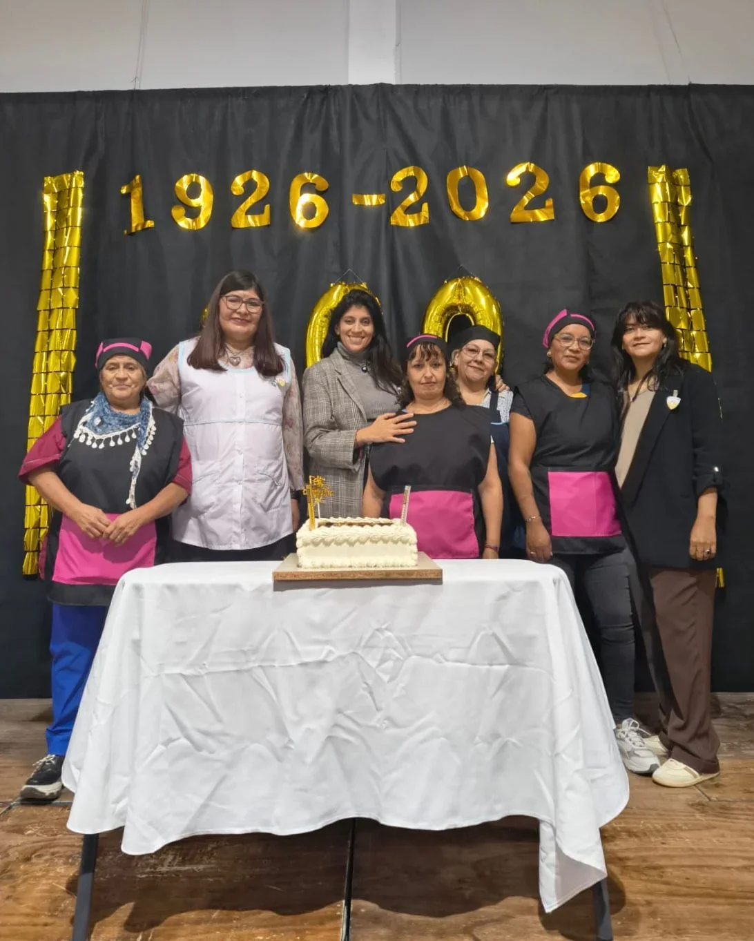 Grupo de mujeres de la Escuela N° 91 junto a una torta frente a un banner que indica el centenario 1926-2026.