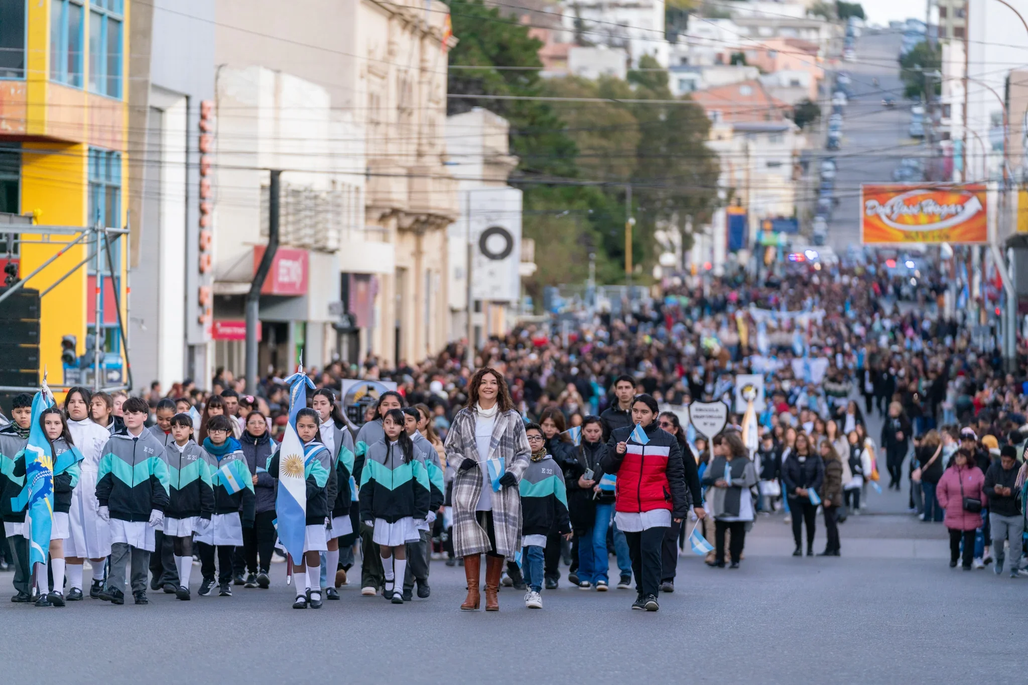 Estudiantes con banderas argentinas y uniformes desfilando en una calle de Comodoro Rivadavia durante la Semana de Mayo.