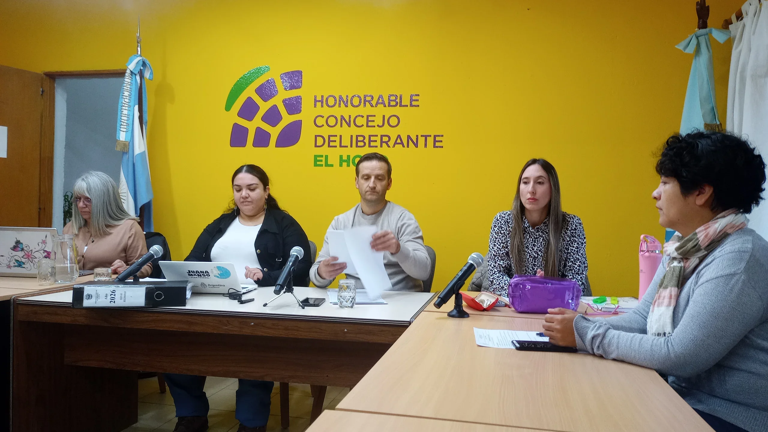 Sesión del Concejo Deliberante de El Hoyo con concejales reunidos frente a un logo institucional sobre fondo amarillo.