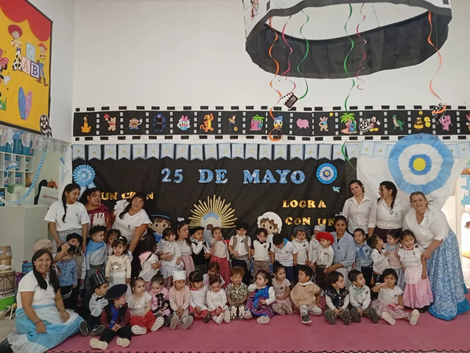 Niños y docentes de un CPI de Puerto Madryn posan frente a un mural del 25 de Mayo con vestimentas típicas y patrias.