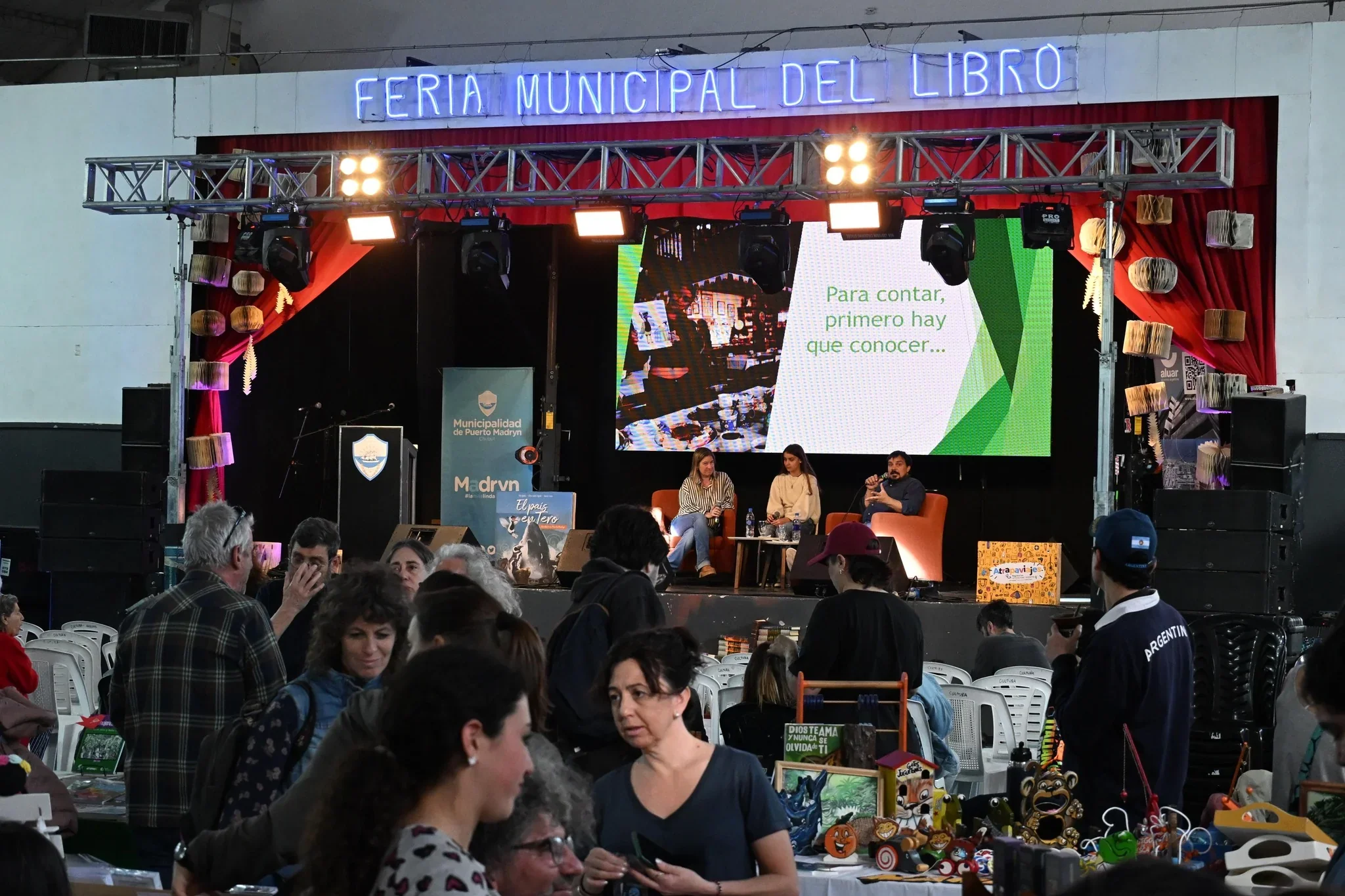 Escenario de la V Feria Municipal del Libro de Puerto Madryn con tres personas conversando y una pantalla gigante.