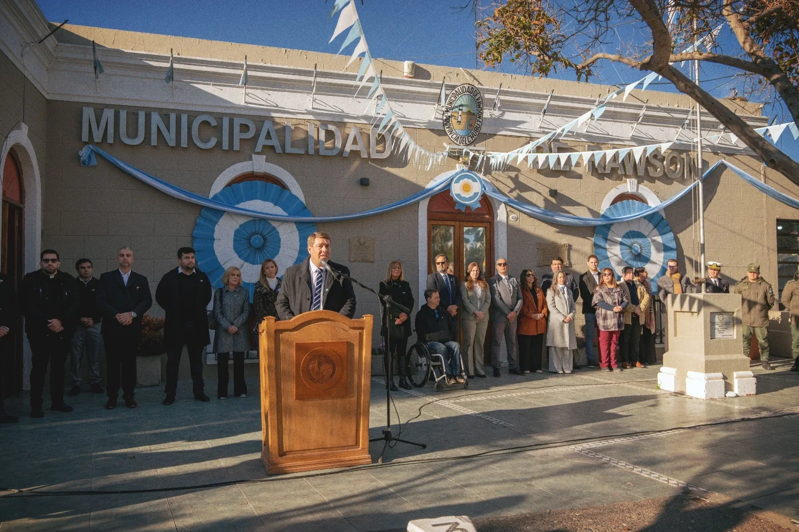 El intendente Damián Biss pronuncia un discurso desde un podio frente a la Municipalidad de Rawson en un acto patrio.