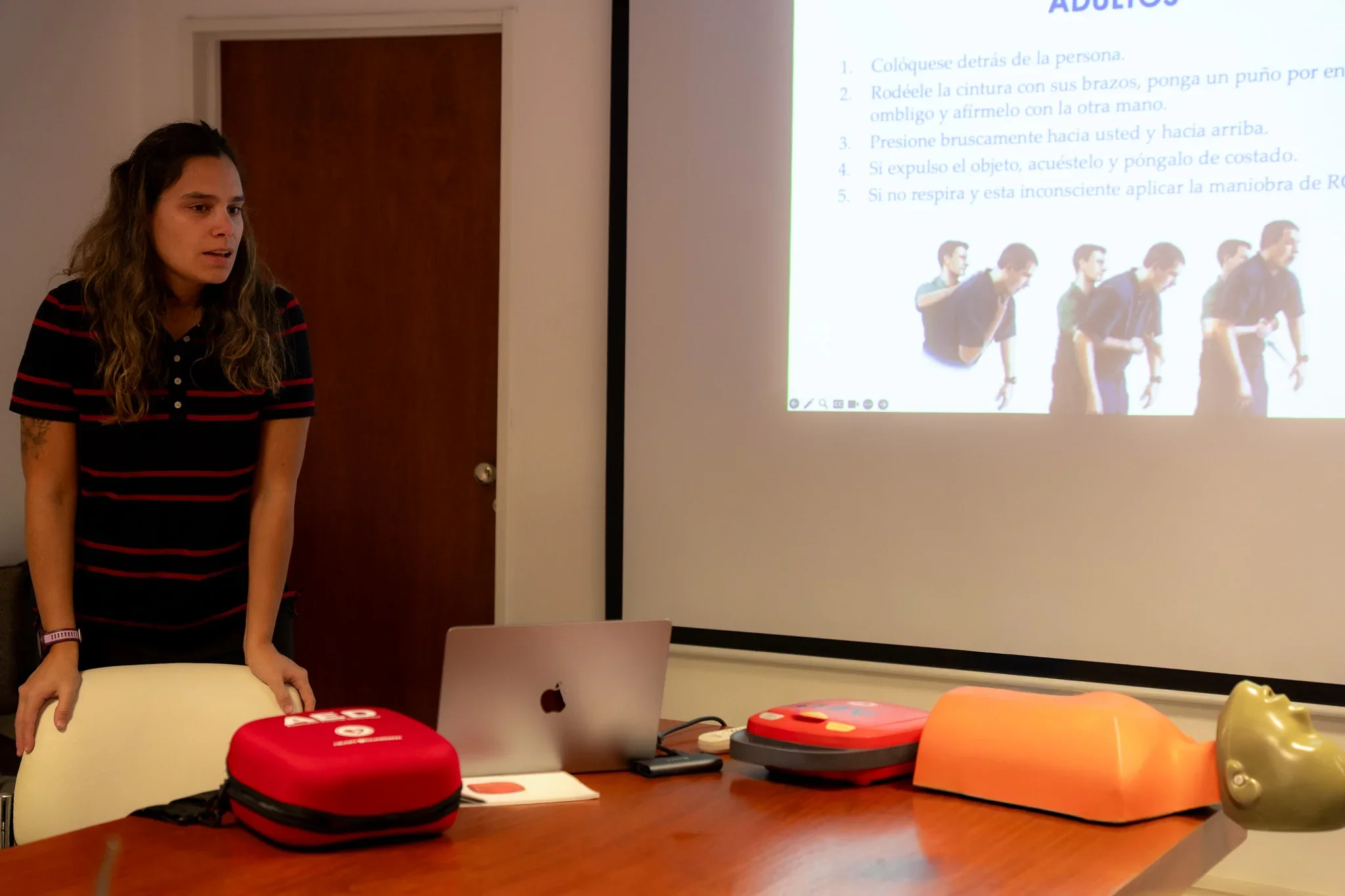 Instructora frente a una proyección sobre primeros auxilios, con un maniquí y un DEA sobre una mesa en Rawson.