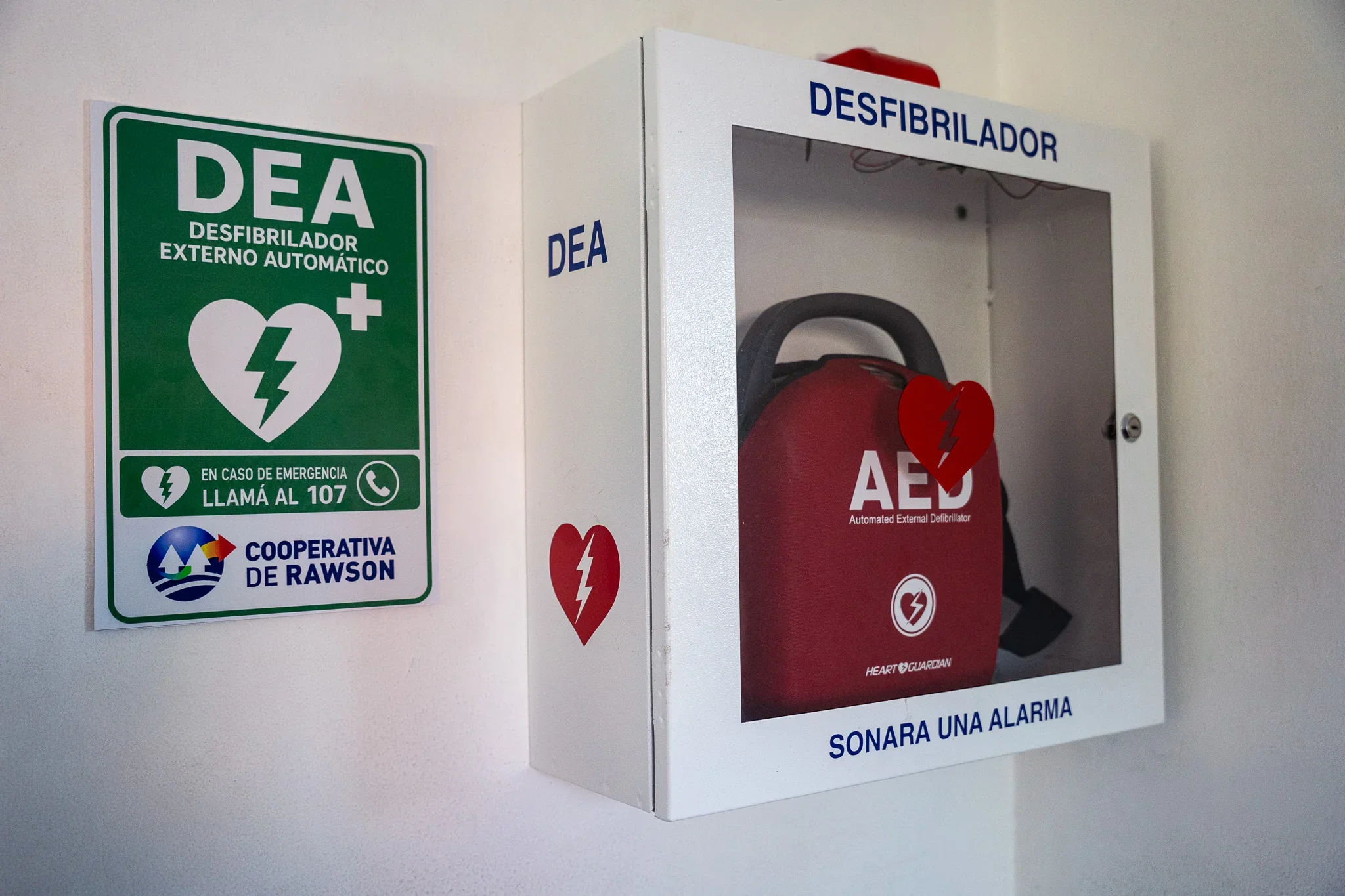 Desfibrilador Externo Automático (DEA) en su caja protectora junto a un cartel informativo de la Cooperativa de Rawson.