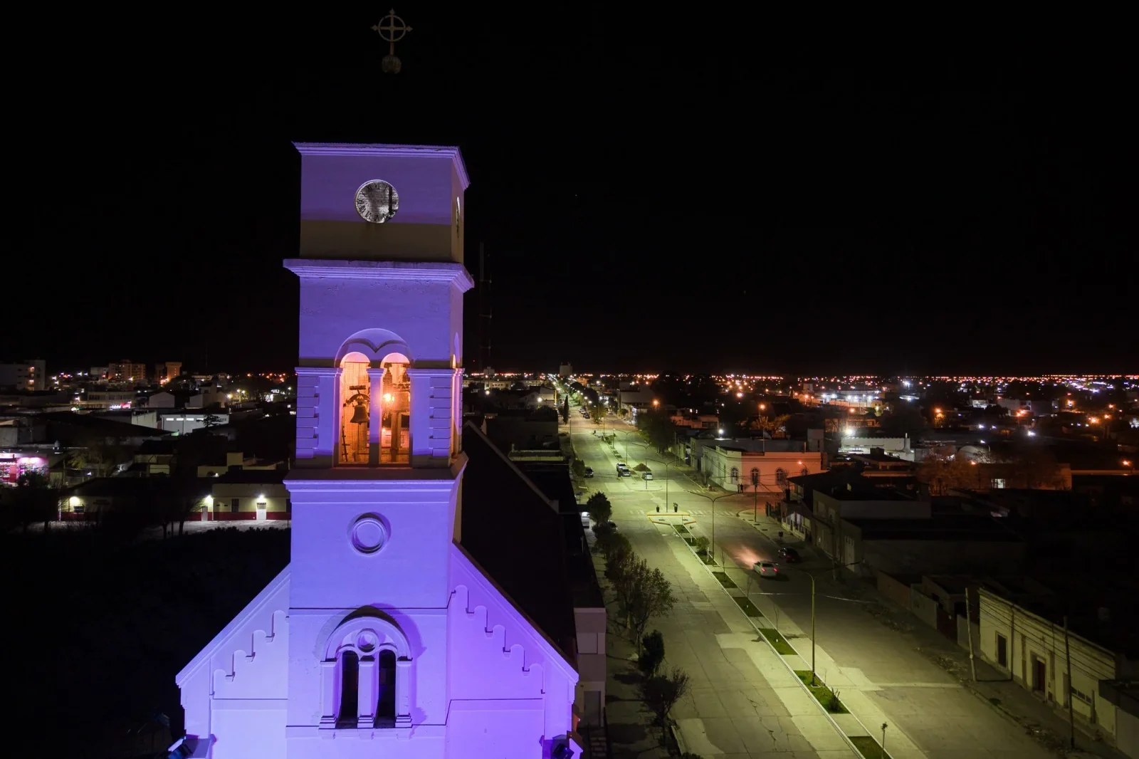 Vista nocturna de la Parroquia María Auxiliadora de Rawson iluminada con luces ornamentales púrpuras en su fachada.