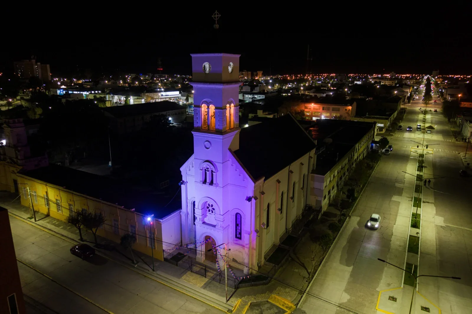 Vista nocturna de la Parroquia María Auxiliadora de Rawson con su fachada iluminada ornamentalmente en color violeta.