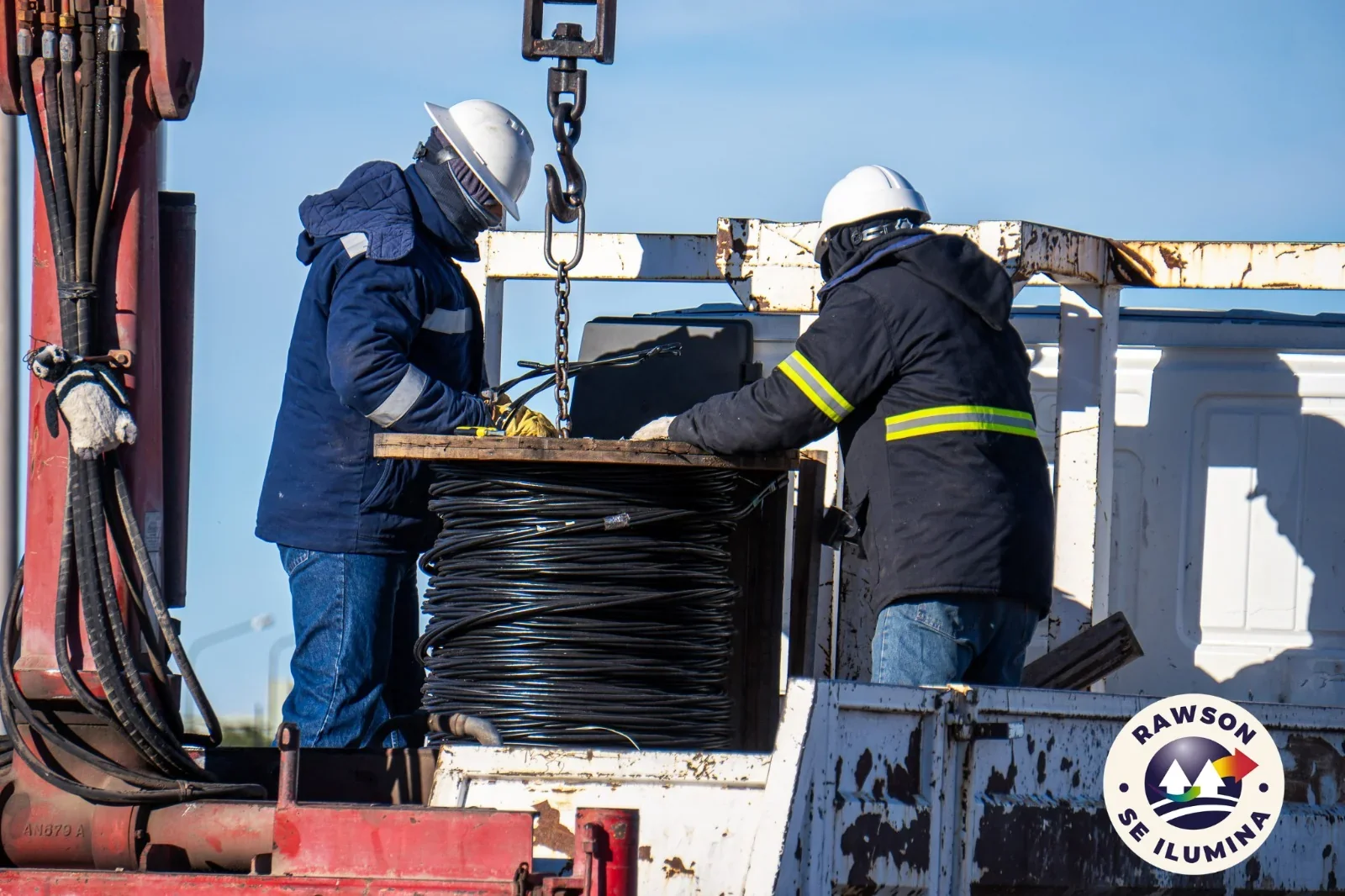 Dos operarios de la Cooperativa con cascos y ropa de seguridad manipulan un rollo de cable eléctrico sobre un camión.