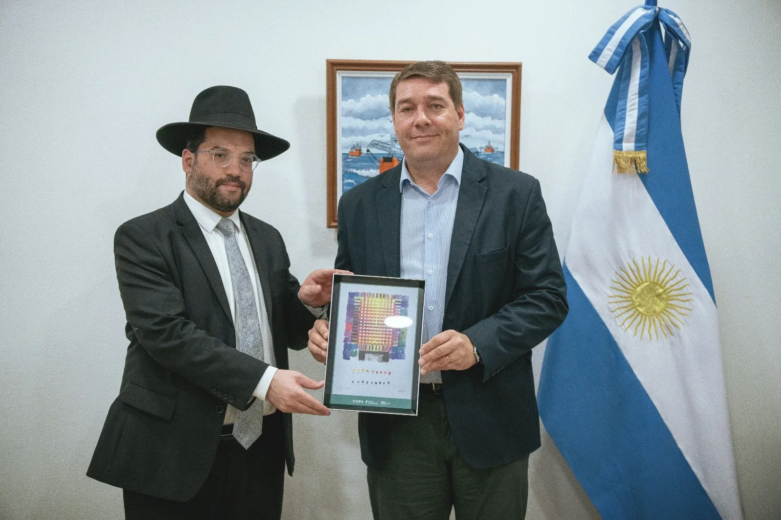 El intendente Damián Biss y el Gran Rabino Eliahu Hamra sosteniendo un cuadro en una oficina con la bandera argentina.