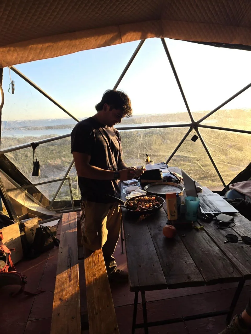 Lautaro Comesaña dentro de un domo geodésico con vista al paisaje costero de Camarones, preparando alimentos.