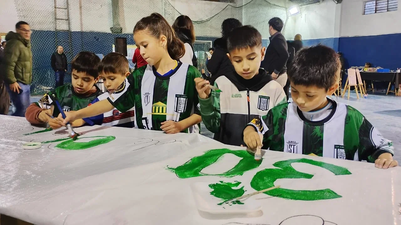 Niños pintando un mural blanco con pintura verde en una actividad cultural comunitaria en Comodoro Rivadavia.