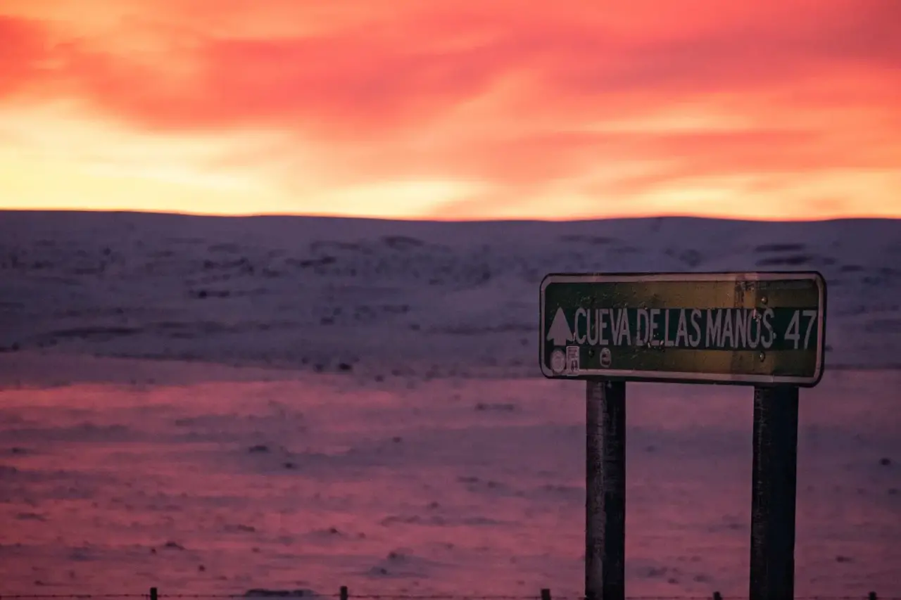 Letrero indicador hacia la Cueva de las Manos en un paisaje patagónico bajo un cielo rojo al atardecer.