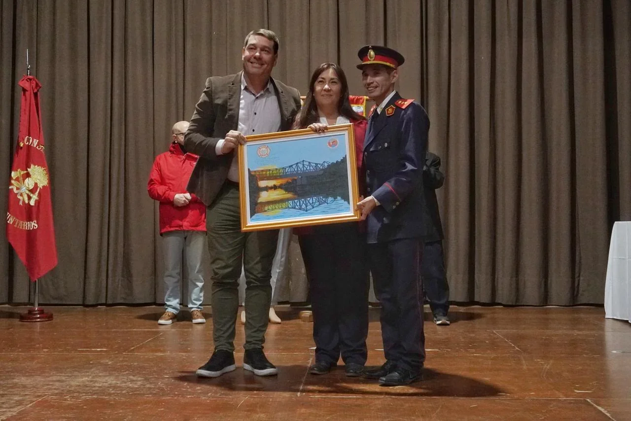 El intendente Damián Biss, Anahí Bustos y un bombero sostienen un cuadro en el 55° aniversario de Bomberos de Rawson.