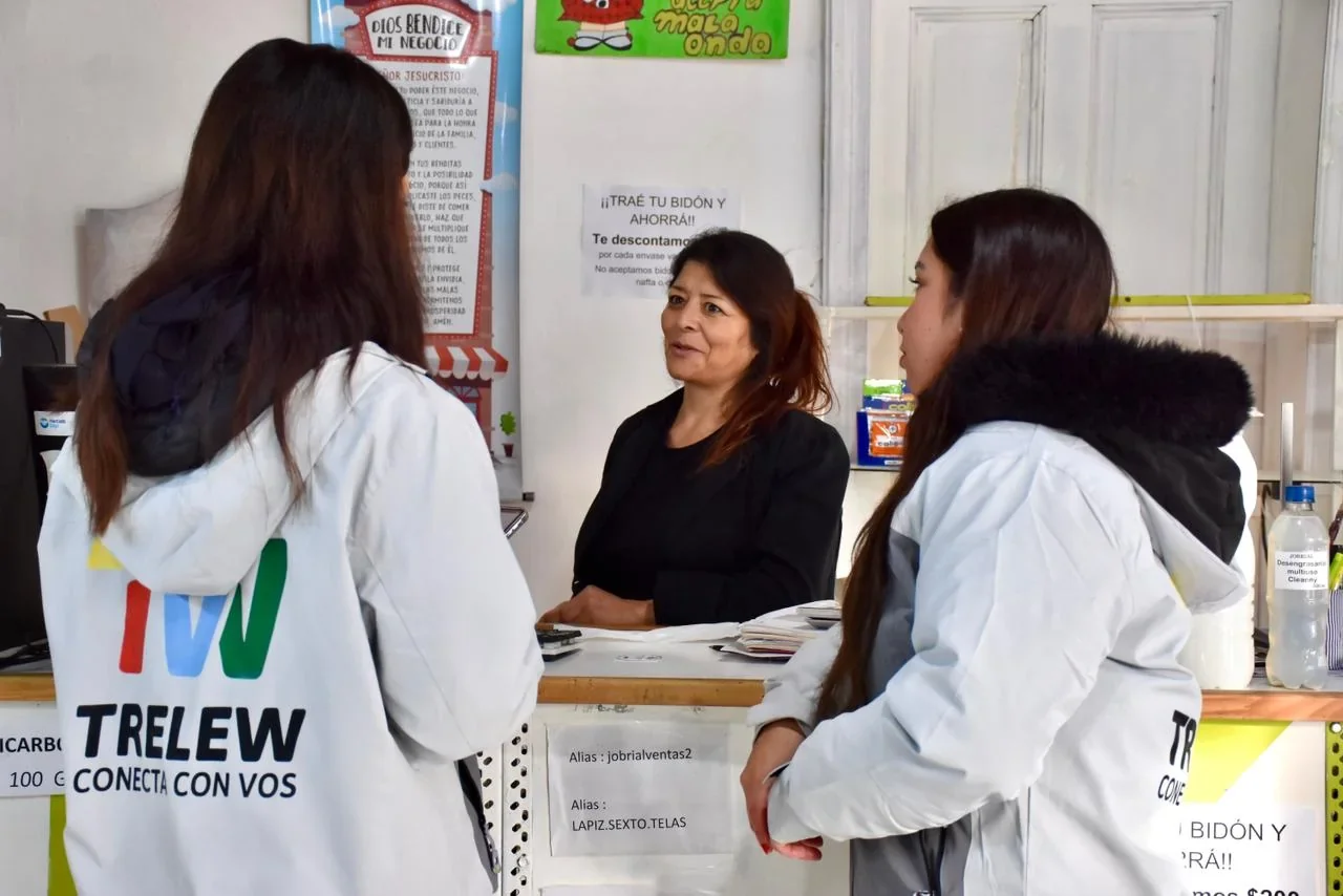 Dos agentes de Atención Ciudadana de Trelew, con camperas blancas institucionales, hablan con una vecina en un comercio.