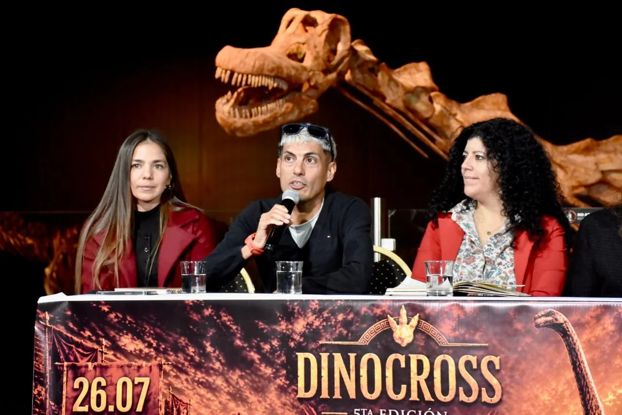 Tres personas en una mesa de presentación de la 5ta edición de la Dino Cross frente a un esqueleto de dinosaurio.